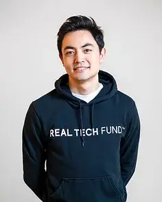 白い壁を背に、REAL TECH FUNDトレーナーを着て、後ろ手に組み笑顔で正面を向く藤井