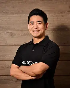 木板貼り合わせを背に、REAL TECH FUNDポロを着て、笑顔で腕を組み左斜めから正面を向く山本